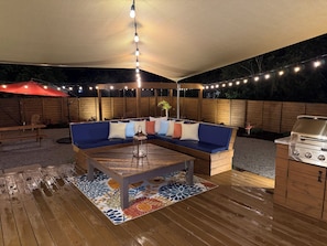 Un patio al aire libre con un sofá azul, una mesa de madera, una parrilla y luces colgantes.