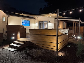 Una terraza de madera con escalones que conducen a un espacio de descanso cubierto, luces colgantes y una pantalla de televisión.