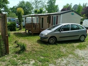 A mobile home with a car parked in front, surrounded by greenery and other mobile homes. - Camping 3 étoiles - Piscine  - ccbgbbc (Thiviers)