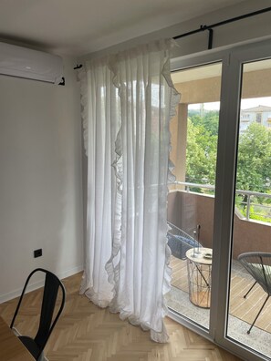 A room with a white curtain, a chair, and a view of a balcony with outdoor furniture.