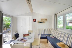 A compact living space with a dining area, a seating area with a striped sofa, and a small table with a magazine and snacks. - Camping 4 étoiles - Piscine  - ccbggc0 (Cordelle)