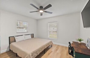 Ein Schlafzimmer mit Bett, Deckenventilator, zwei Fenstern mit Jalousien, Schreibtisch mit Stuhl und einer Pflanze.