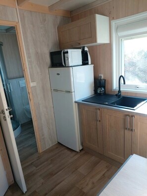 A compact kitchen with a refrigerator, microwave, and coffee maker, wooden cabinets, a window with a view, and a door leading to a bathroom.