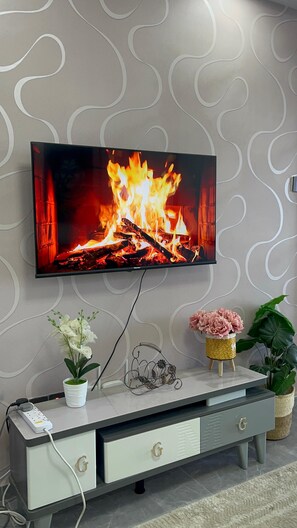 Et tv ophængt på en væg viser et bål, en hvid potteplante, en dekorativ metaldel og en lille fletkurv med pink blomster.