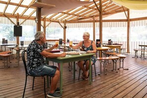 An outdoor dining area with wooden tables and chairs, covered by a wooden roof structure. - Camping 3 étoiles - Piscine  - ccbfgda (Les Achards)