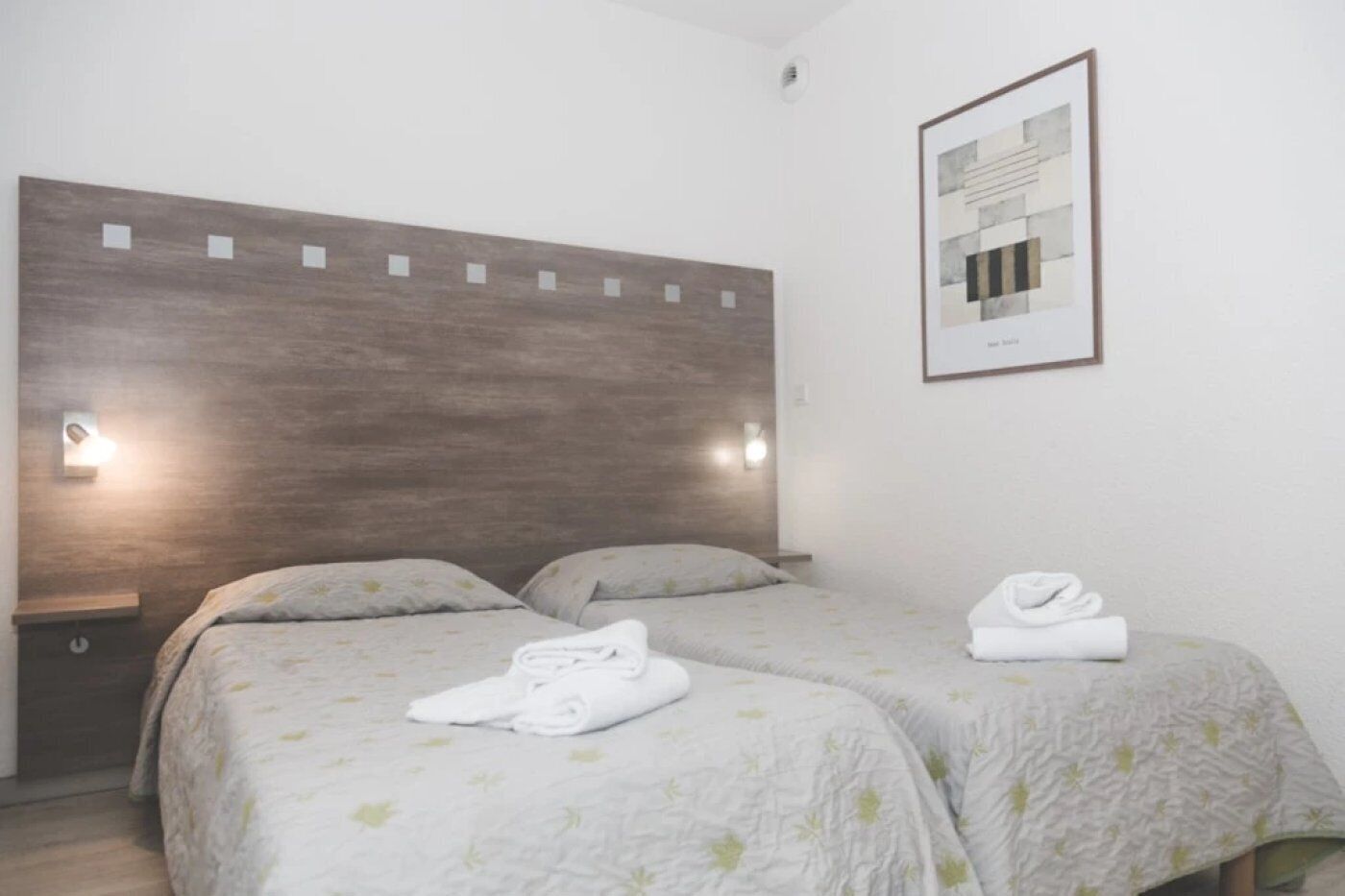 Een hotelkamer met twee bedden, een houten hoofdbord, wandlampen en een lijst met een afbeelding.