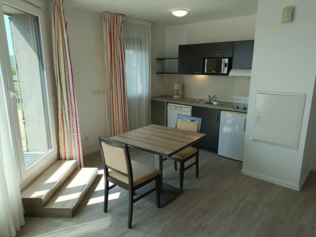 Een kleine keuken met een magnetron, spoelbak en vaatwasser. Er is een eettafel met stoelen en een raam met gordijnen.