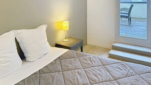 Quarto com cama, mesa de cabeceira com abajur e janela com vista para uma cadeira e um corpo d'água.