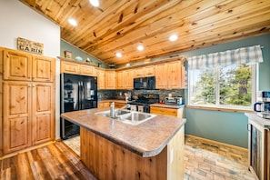 A kitchen with wooden cabinets, a granite countertop, a stainless steel sink, a refrigerator, a microwave, and a window with a view of trees. - The Office With Hot Tub and Dogs OK (Duck Creek Village)