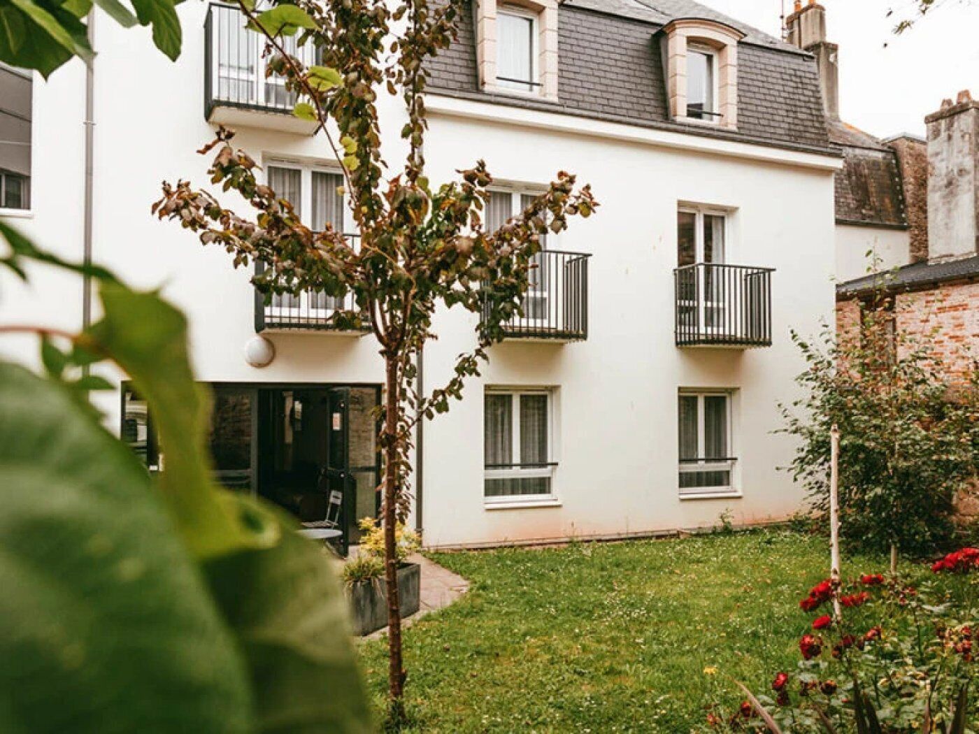 Un edificio blanco con múltiples ventanas y balcones, rodeado de un espacio verde con plantas y flores.