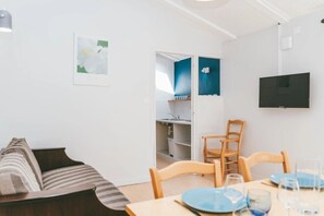 A room with a sofa, a dining table set with plates and glasses, a TV mounted on the wall, and a kitchen area visible through an open door. - Camping 2 étoiles - Piscine  - ccbgfed (Pluherlin)