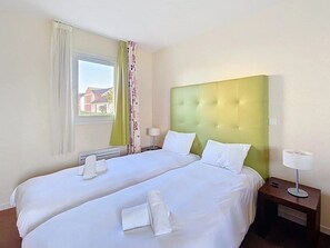 Une chambre d’hôtel avec deux lits, une fenêtre avec des rideaux, une table de chevet avec une lampe, et une vue sur les bâtiments à l’extérieur.