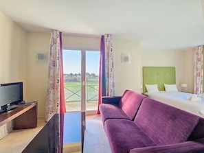 A hotel room with a television, a sofa, a bed, and a view of a green landscape through the window. - Appartement 3 étoiles - Piscine  - ccbgeeh (Amboise)