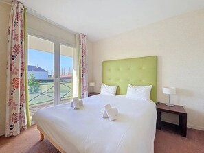 Een hotelkamer met een groot bed, een groen hoofdeinde, een nachtkastje met een lamp, en uitzicht op een gebouw door het raam.