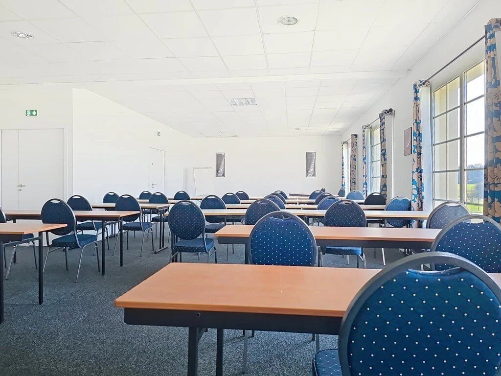 Ein Konferenzraum mit Tischen und Stühlen, einer weißen Decke und Fenstern mit Vorhängen.