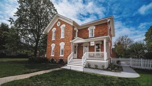 A two-story brick house with a front porch, white railings, and a small garden. - Charming Restored Historic 3BR Home - The Larrabee (Williamston)