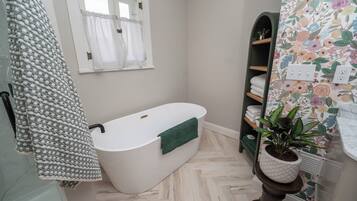 A bathroom with a freestanding bathtub, a window with curtains, a shelf with towels, and a potted plant.