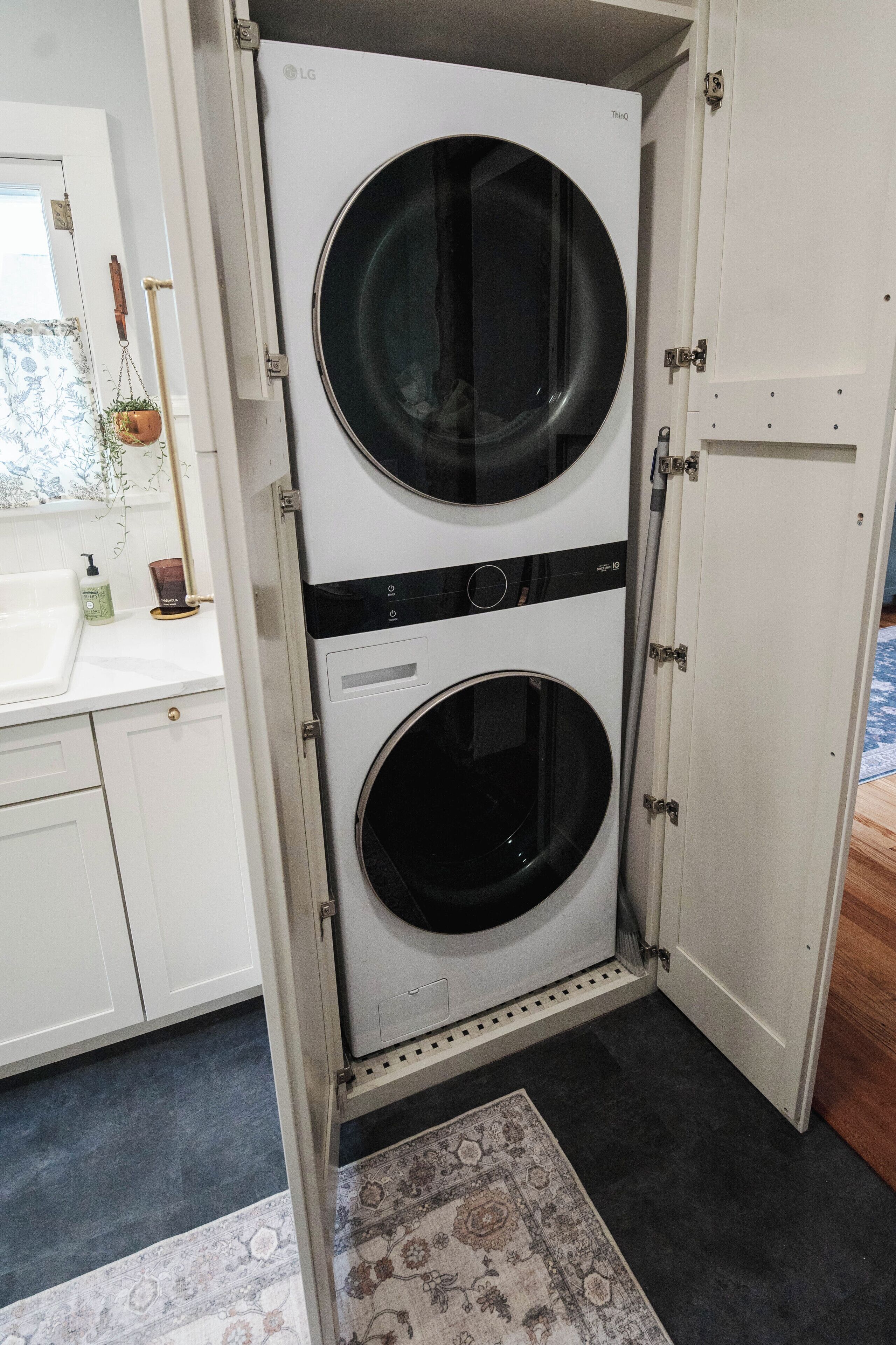 A compact, white laundry room with two stacked front-loading washing machines, a small sink, and a decorative rug.