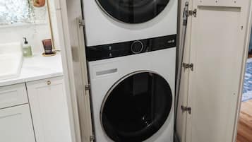 A compact, white laundry room with two stacked front-loading washing machines, a small sink, and a decorative rug.