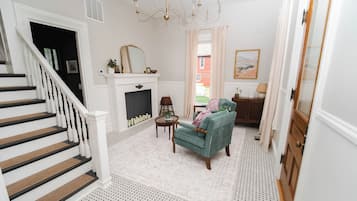 Un escalier avec des balustrades blanches, une cheminée surmontée d’un miroir, un fauteuil turquoise et un tapis à motifs.