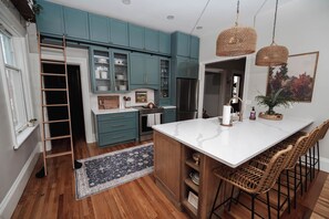 Une cuisine avec des meubles de couleur turquoise, un ßlot central blanc, des chaises en osier et une échelle.