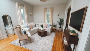 A living room with a sofa, armchair, coffee table, and a large mirror. - Charming Restored Historic 3BR Home - The Larrabee (Williamston)