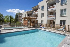 A swimming pool with lounge chairs, a wooden pergola, and a multi-story building with balconies. - Appartement 3 étoiles - Piscine  - ccbgfcd (Gréoux-les-Bains)