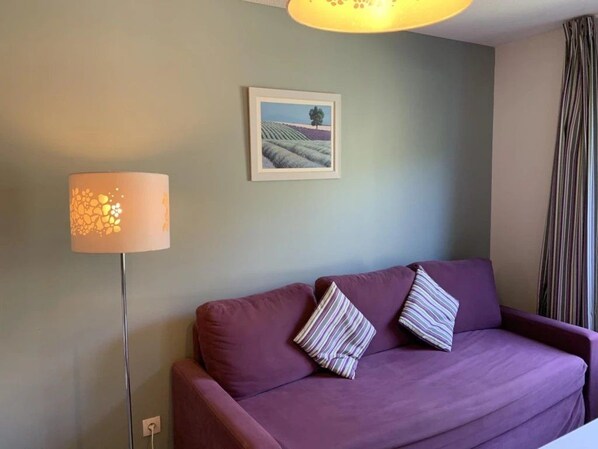 A room with a purple sofa, two striped pillows, a floor lamp, and a framed picture on the wall.