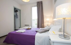 A hotel room with a bed, a bedside table, a lamp, a window with curtains, a door, and a small balcony.
