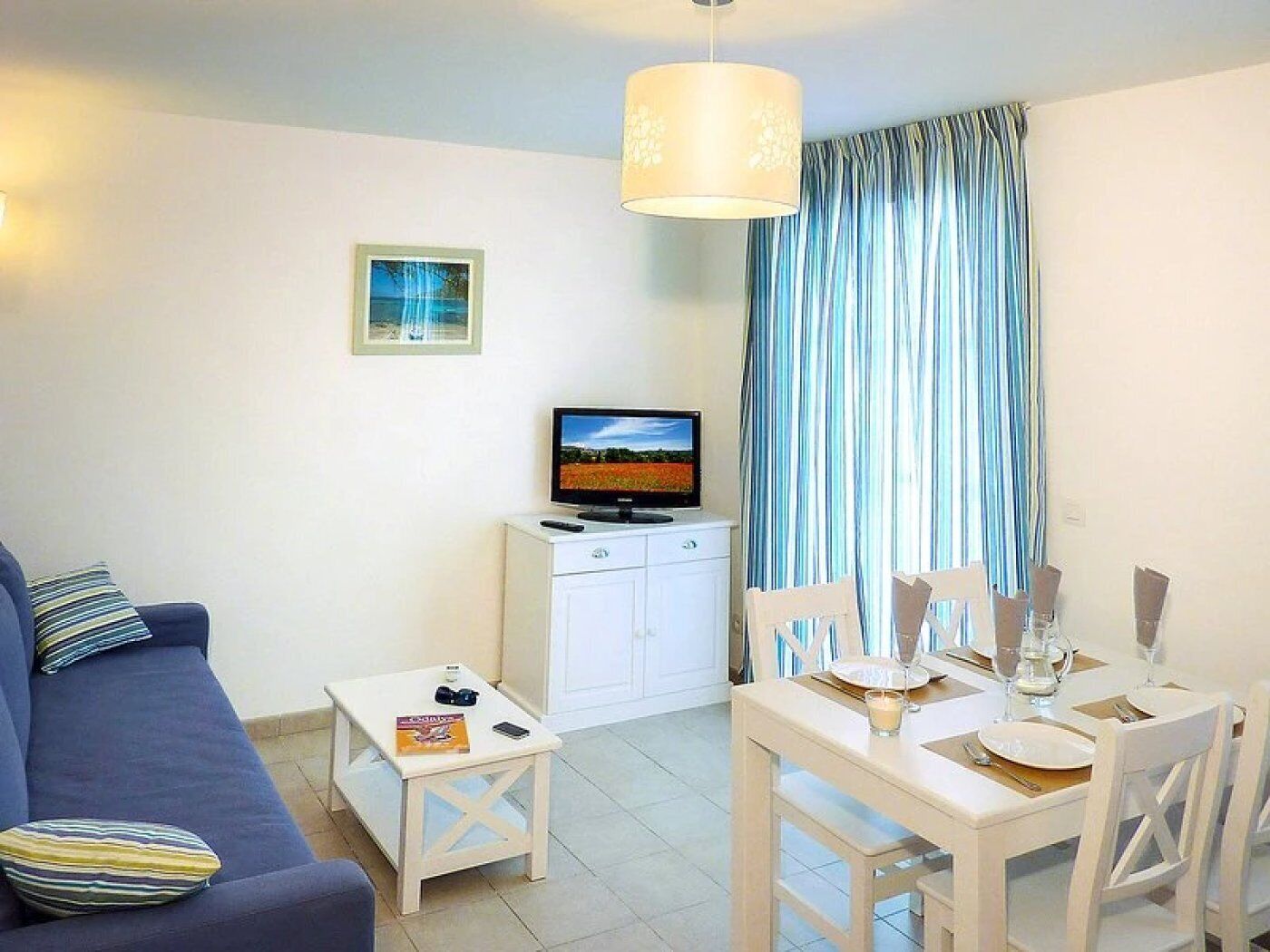 A living room with a sofa, a TV, a dining table set for two, and a window with curtains.