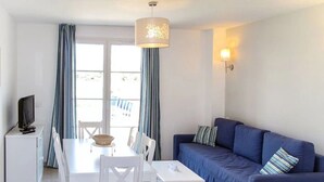 A room with a dining table, chairs, a blue sofa, a television on a cabinet, and curtains. - Camping 3 étoiles - ccbgfbi (Cabriès)
