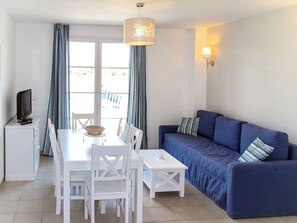 Ein Zimmer mit Esstisch, Stühlen, einem blauen Sofa, einem Fernseher auf einem Schrank und Vorhängen.