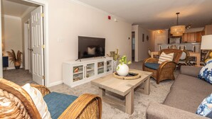 A living room with a television, wicker furniture, a coffee table, and a kitchen in the background. - Rehoboth Perfect 2BR Condo sleeps 6 with a Pool (Rehoboth Beach)