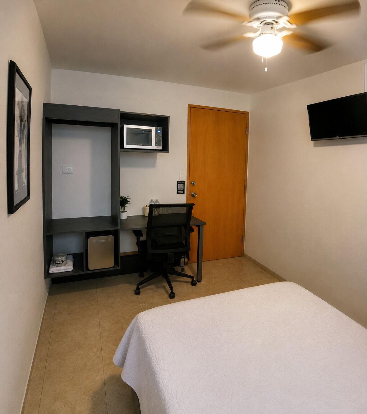 A bedroom with a bed, a desk with a chair, a microwave, a door, and a ceiling fan.