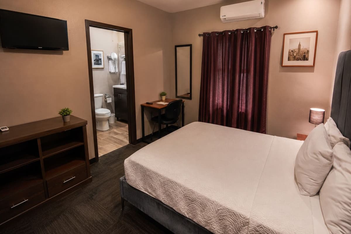 A hotel room with a bed, a desk, a chair, a TV, a mirror, a bathroom, and an air conditioner.