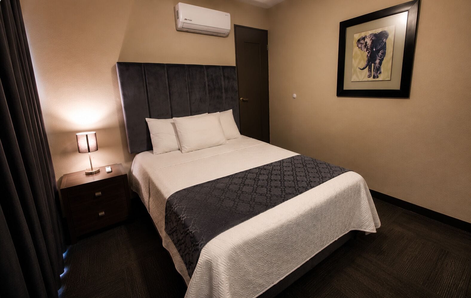 Ein Hotelzimmer mit Bett, Nachttisch, Lampe, Klimaanlage und einem gerahmten Bild an der Wand.
