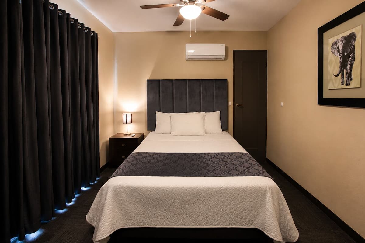 A hotel room with a bed, a nightstand, a ceiling fan, an air conditioning unit, and a framed picture on the wall.