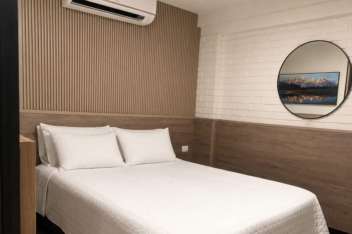 A bed with a white quilted bedspread, a wooden headboard, and a white brick wall with a round mirror reflecting a mountain landscape.