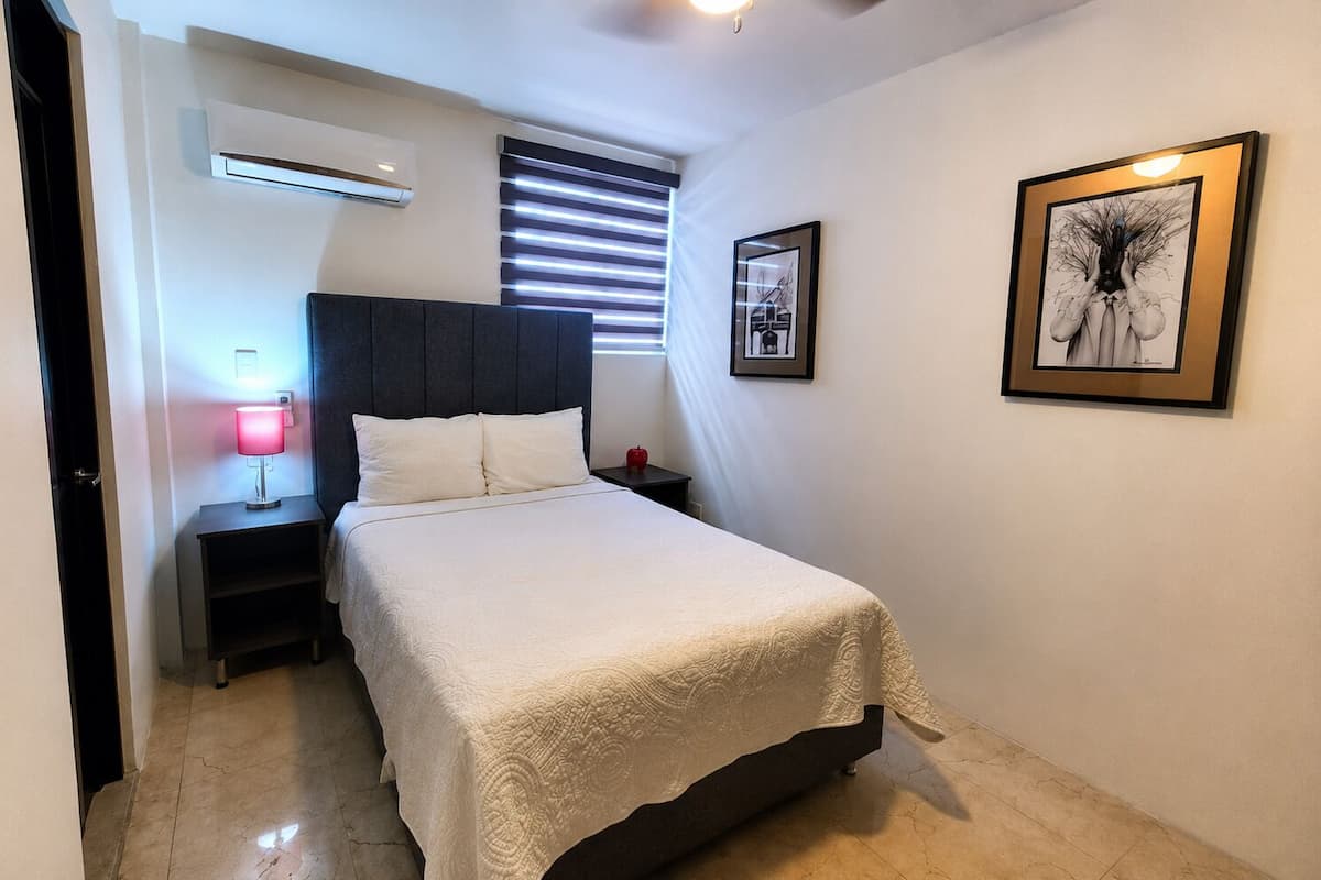 A bedroom with a bed, bedside tables, a lamp, an air conditioner, and two framed pictures on the wall.