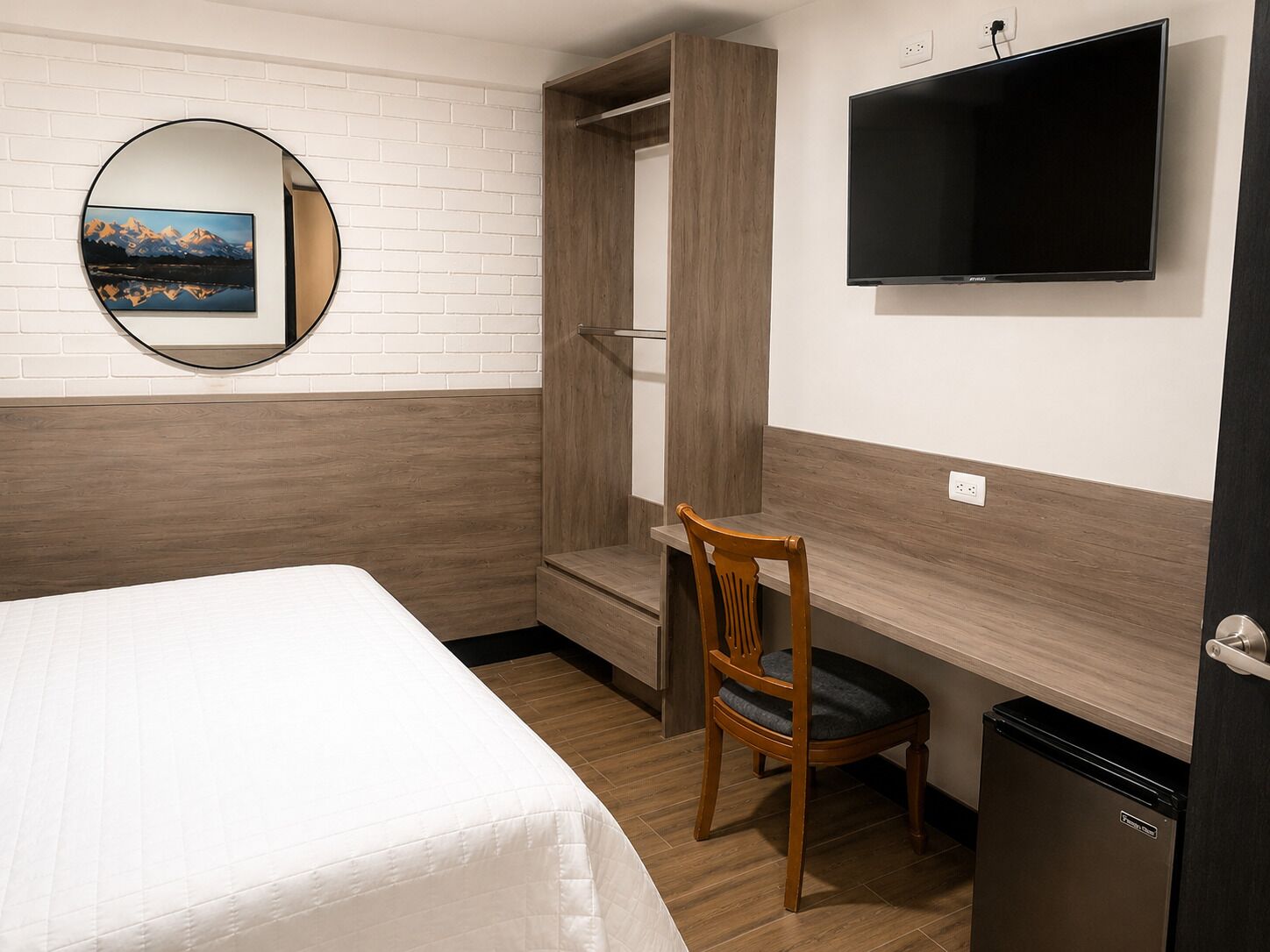 A hotel room with a bed, a desk with a chair, a television, and a compact refrigerator.