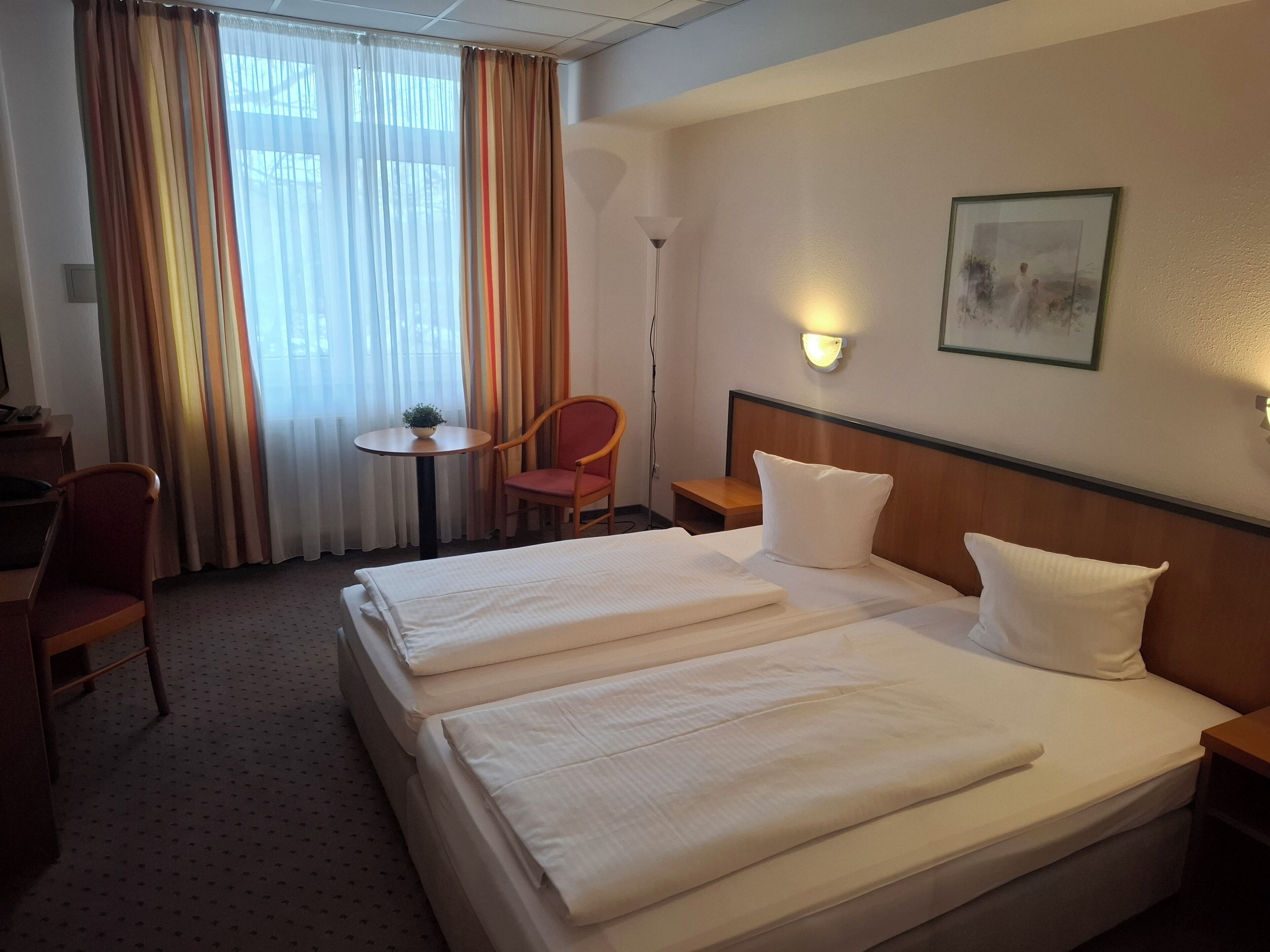Une chambre d’hôtel avec deux lits, un bureau, une chaise, une petite table avec une plante et une lampe fixée au mur.