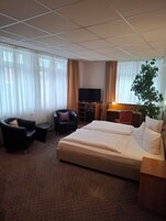 Habitación de hotel con una cama, dos sillones, un televisor, un escritorio y una planta.