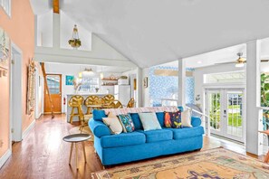 A spacious living room with a blue sofa, wooden dining table, and chairs, a kitchen in the background, and large windows.