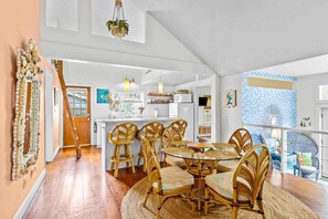 A spacious dining area with a round table and wicker chairs, a kitchen in the background, and a staircase leading to another level. - The Mermaid Cottage - Walk to Waterfront (Morehead City)