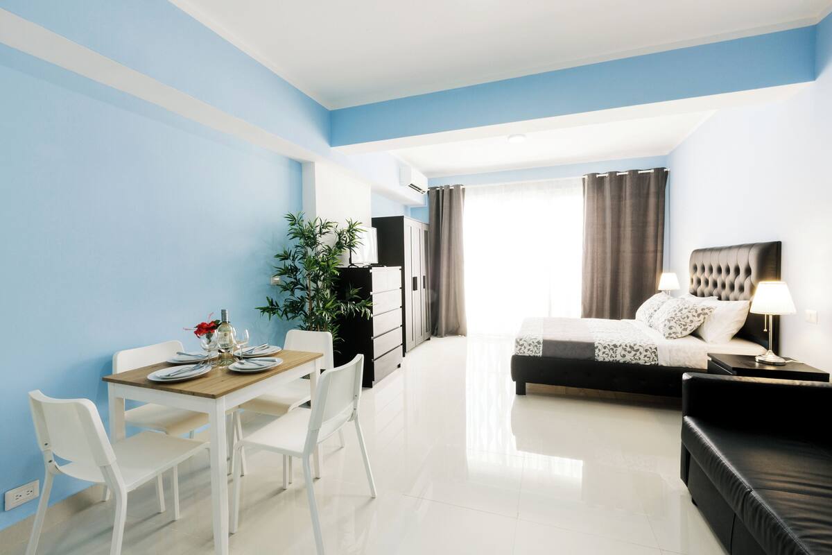 A modern hotel room with a bed, bedside table, and a dining area with chairs and a table.