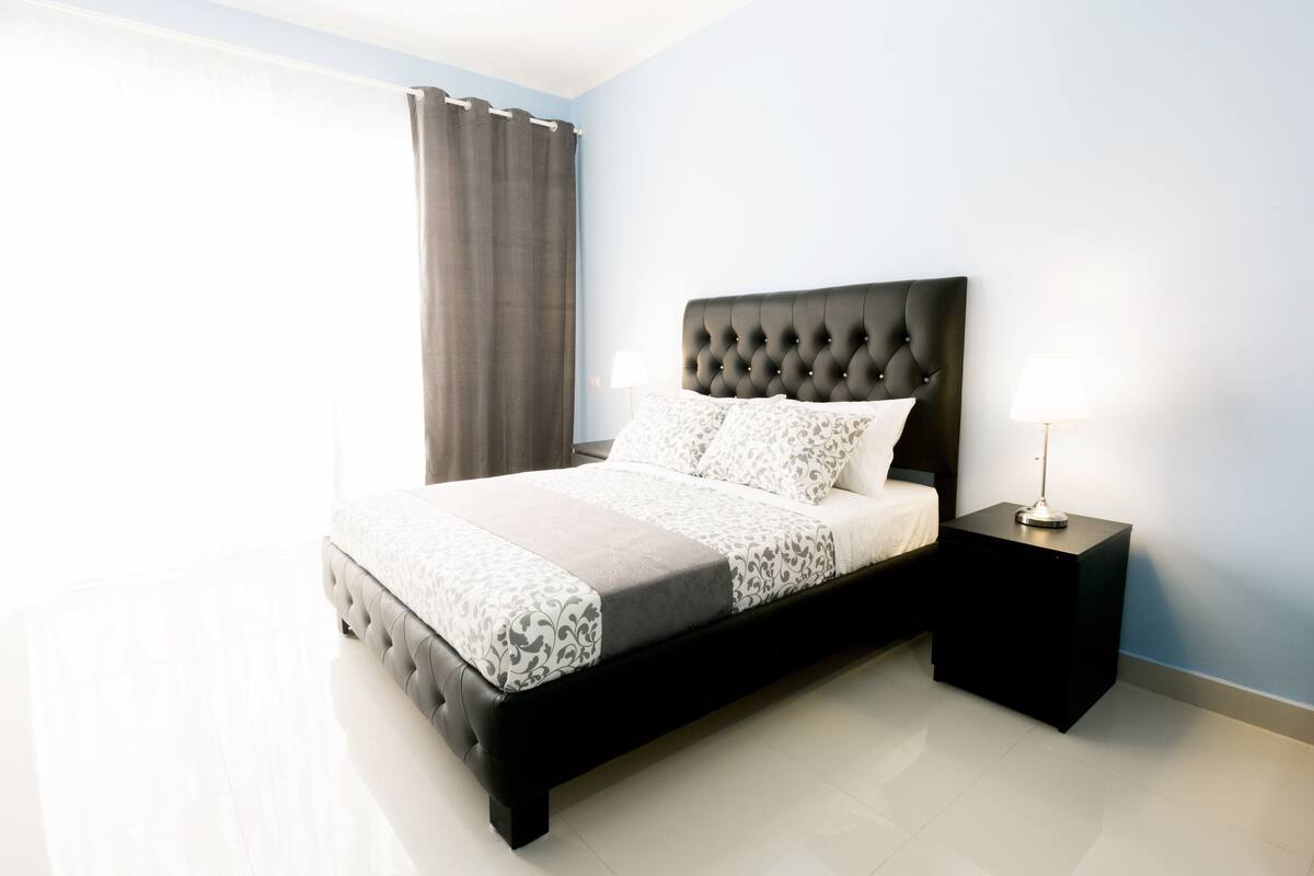 A modern bedroom with a large bed, a nightstand, a lamp, and a window with curtains.