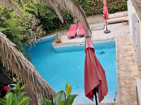 Une piscine dotée d’un toit de chaume, entourée d’une végétation luxuriante et agrémentée d’un espace détente avec des fauteuils roses.