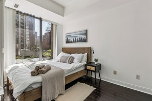 Un dormitorio con cama, ventana, mesita de noche y un cuadro colgado en la pared.