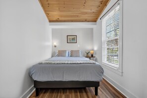 Ein Schlafzimmer mit Bett, Holzboden, weißen Wänden, einem Fenster mit Jalousien, einem Nachttisch mit Lampe und einem gerahmten Bild an der Wand.