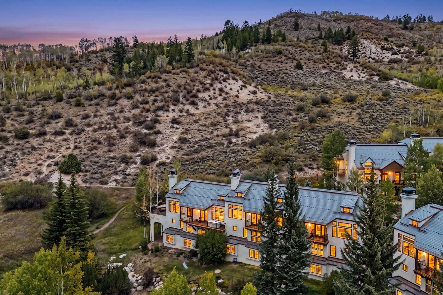 A large, multi-story house with a modern design, surrounded by a natural landscape with trees and hills.